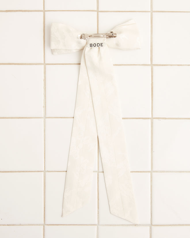 The Football Jacquard Bow Barrette in cream, which channels the vintage style of the 1950s-1960s with its long tails, is displayed on a white tiled background.