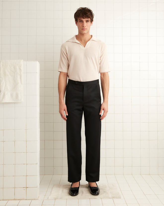 In a minimalist, tiled room, a person stands wearing a light shirt and the sleek Barathea Tuxedo Trousers with satin tuxedo stripes, hands resting at their sides.