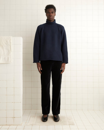 A person dressed in a dark blue sweater, a pair of Alpineflower Trousers, and shoes stands against a white tiled wall in a minimalist setting.