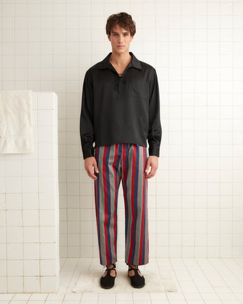 A person in Marina Stripe Pajama Pants and a blazer stands next to a door, leaning against the frame with one hand, in an outdoor setting with a lounge chair nearby. The classic pajama silhouette of their attire evokes both comfort and timeless style.