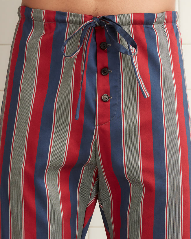 A person in Marina Stripe Pajama Pants and a blazer stands next to a door, leaning against the frame with one hand, in an outdoor setting with a lounge chair nearby. The classic pajama silhouette of their attire evokes both comfort and timeless style.