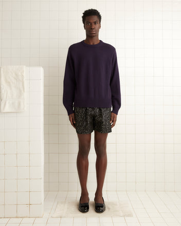 A person wearing Glitter Shorts and a purple sweater, embellished with subtle glitter dots, stands in a white tiled room.