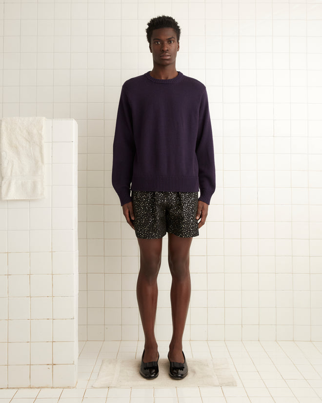 A person wearing Glitter Shorts and a purple sweater, embellished with subtle glitter dots, stands in a white tiled room.