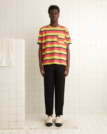 A person wearing a Rec Stripe Pocket Tee, reminiscent of 1980s style, along with black pants, stands in a tiled room.