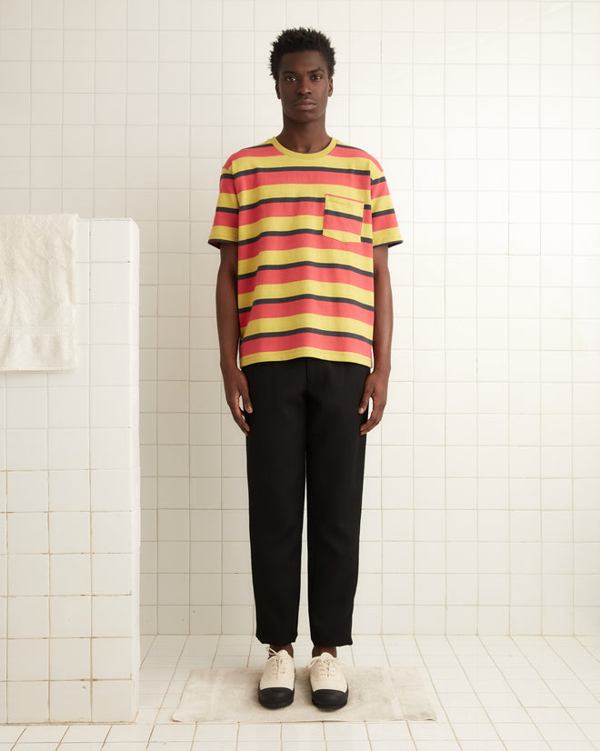 A person in a Rec Stripe Pocket Tee and black pants stands in a tiled room with a towel hanging nearby.