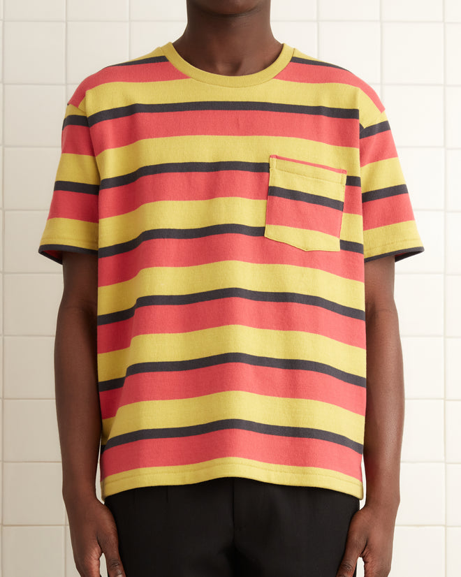 A person in a Rec Stripe Pocket Tee and black pants stands in a tiled room with a towel hanging nearby.