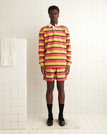 A person wearing a striped yellow and red outfit stands on a white tiled floor, with black socks and shoes, completed by Rec Stripe Sweatshorts for a retro 1980s-inspired look.