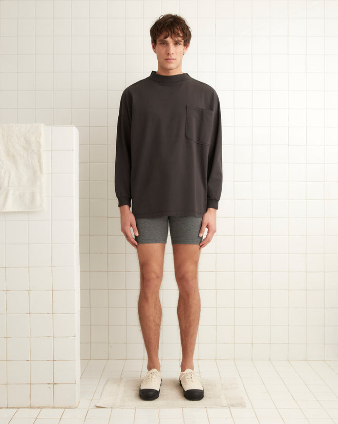 A person stands in a tiled room, wearing a Barry Mockneck dark long-sleeve shirt made from midweight cotton, gray shorts, and beige shoes, facing the camera with a neutral expression.