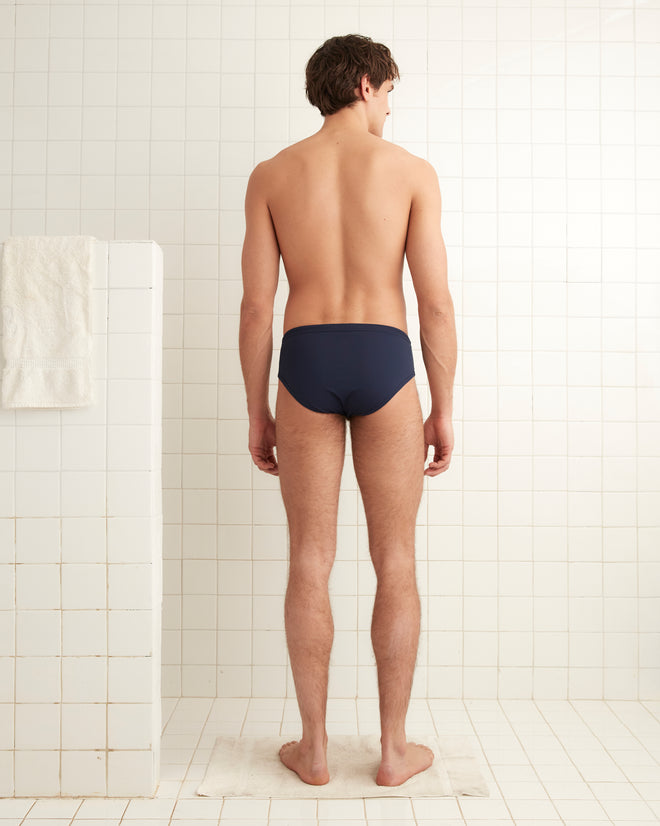 A man wearing Bode's Navy Swimming Briefs, with a discreet logo, faces away in a tiled shower area.