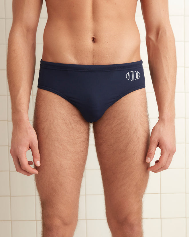 A man stands against a tiled wall wearing Swimming Briefs - Navy, featuring the Bode logo.