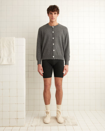 A person wearing a gray cardigan crafted from luxurious Royal Alpaca and Lounge Shorts in black stands gracefully in a white tiled room.