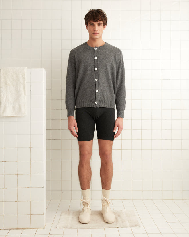 A person in a gray cardigan and black Lounge Shorts stands in a tiled room, showcasing the comfort of beige socks and light shoes made from organic cotton.