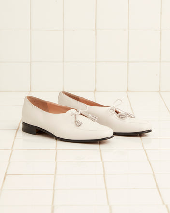 The Tassel Shoes - White, a pair of vintage white leather loafers with tassels on top, made in Italy, is placed on a tiled floor.