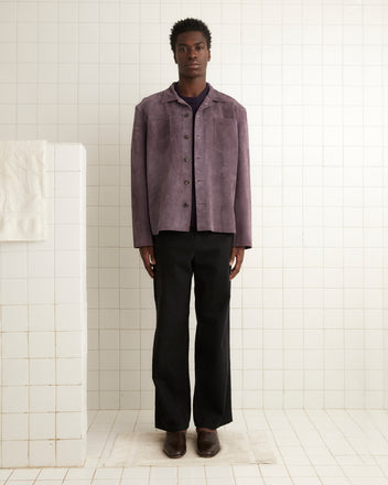 A person wearing a purple Suede Overshirt and dark pants stands in a tiled room with a towel on a rail.