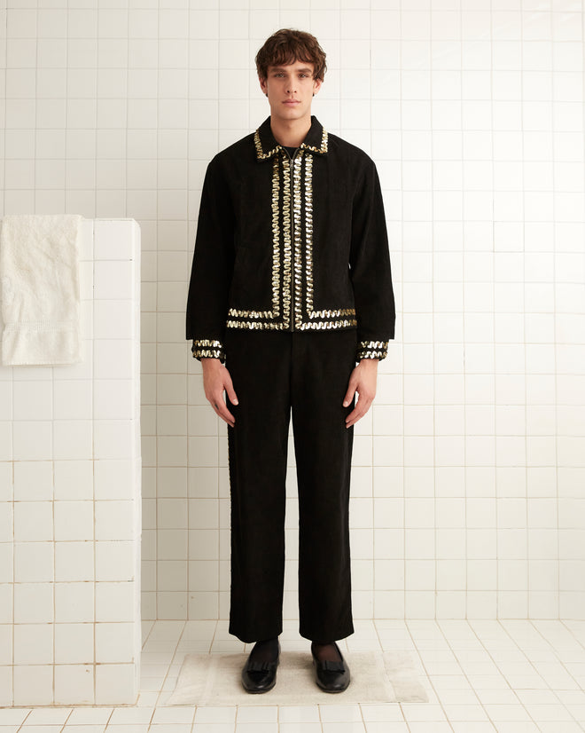 A person stands in a tiled room, wearing the Bandstand Jacket in black with gold sequin trim, black shoes, and looking directly ahead.