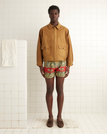 A person stands in a white tiled room wearing a Coach's Jacket - Tan, colorful shorts, and brown slippers, with a towel hanging on a nearby white partition.