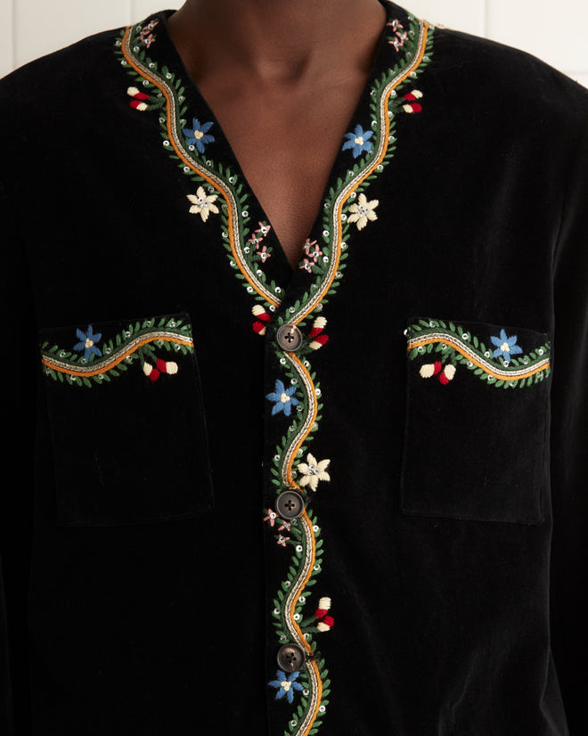 Person in an Alpineflower Jacket, featuring vintage-inspired floral embroidery, stands in a tiled room with a towel on the towel rack.