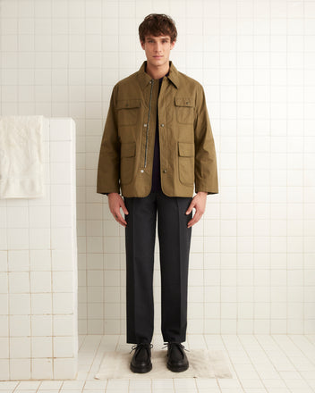 A man stands in a tiled room wearing a brown Waxed Hunting Jacket, a black shirt, dark trousers, and black shoes.