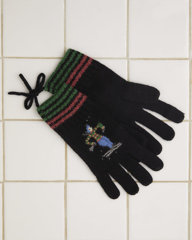 The Ski Run Gloves, made from merino wool and featuring black knit with red and green stripes, showcase a small embroidered character, elegantly displayed on a tiled surface.