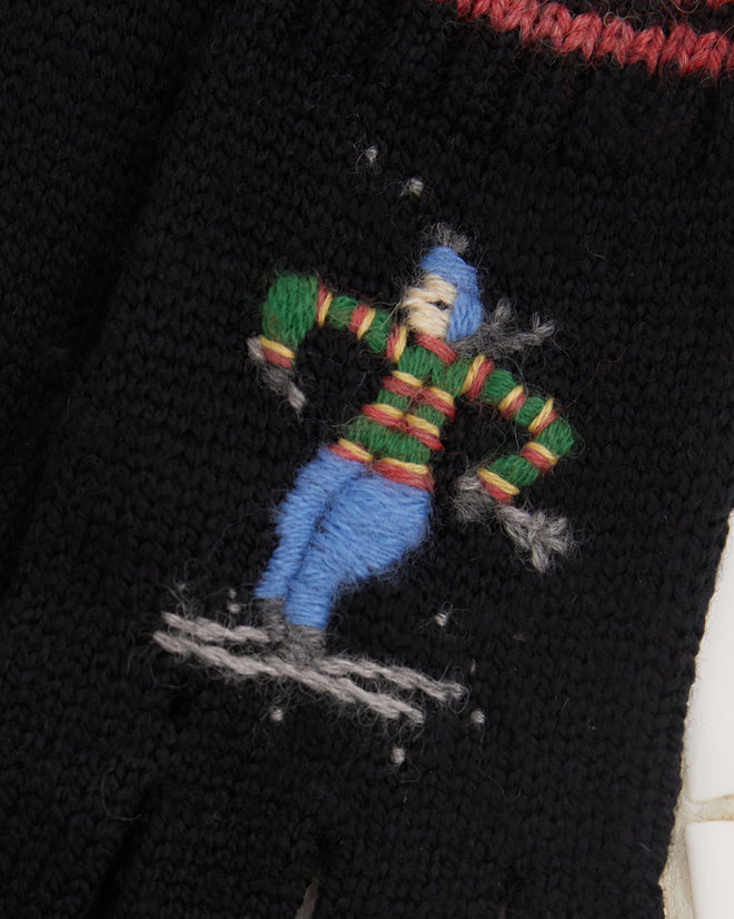 The Ski Run Gloves, made from merino wool and featuring black knit with red and green stripes, showcase a small embroidered character, elegantly displayed on a tiled surface.