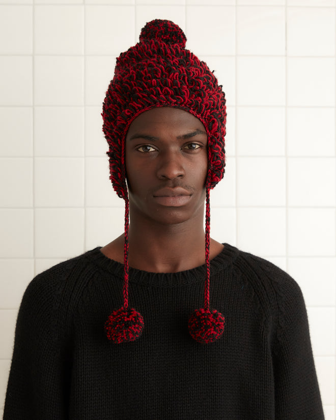 The Loopy Tassel Hat, expertly crocheted from soft merino wool in a fuzzy red and black design with playful pom-pom tassels, is showcased against a crisp white tiled background.