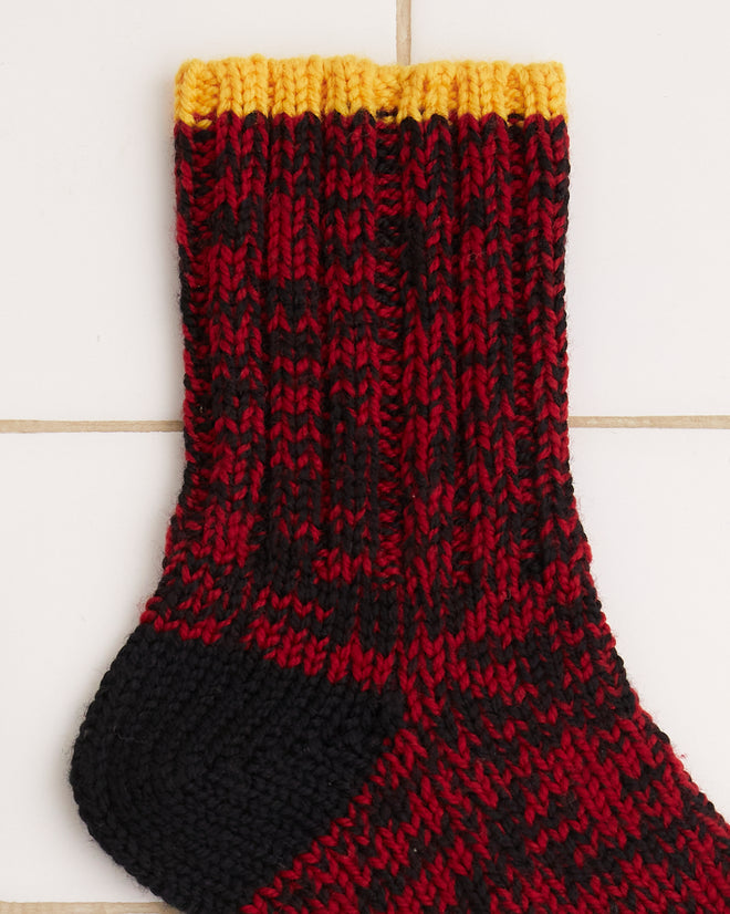 Two Melange Socks with a red and black speckled design and yellow accents, reminiscent of merino wool patterns, rest on a white tiled background.