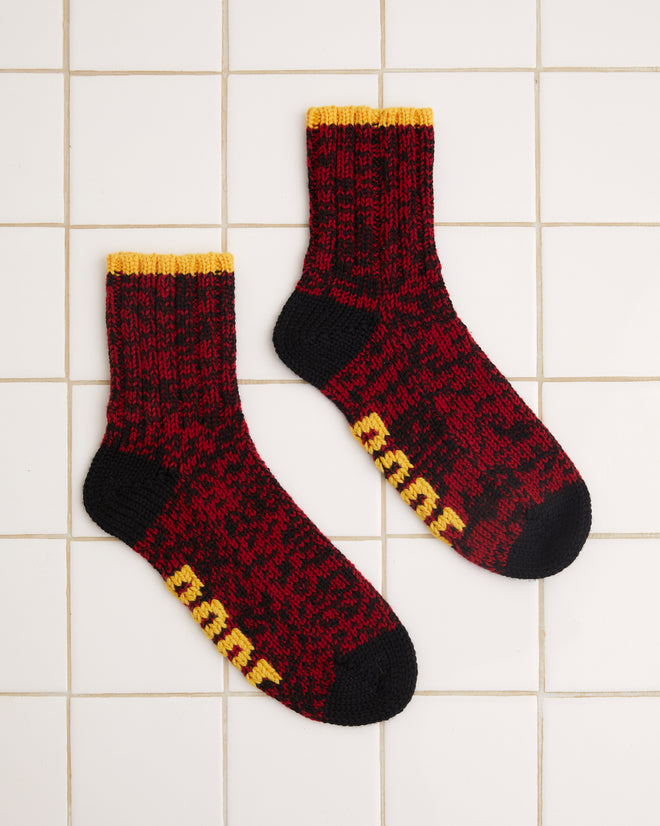 Two Melange Socks with a red and black speckled design and yellow accents, reminiscent of merino wool patterns, rest on a white tiled background.