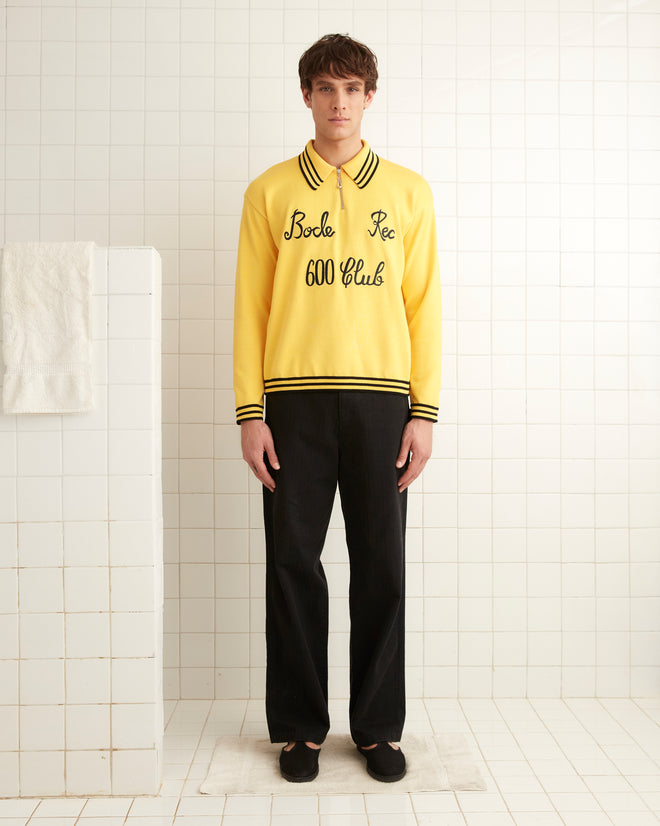 A person in a cozy, Club Long-Sleeved Polo - Yellow and blue jeans sits on a lounge chair in front of a wall with signs for dressing rooms and emergency information.