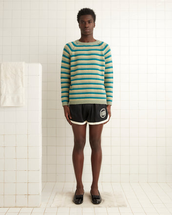 In a tiled room, an individual confidently sports the Mossy Glen Crewneck, a striped 100% wool sweater, paired with black shorts and shoes.