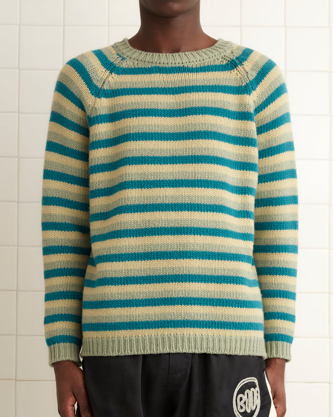 In a tiled room, an individual confidently sports the Mossy Glen Crewneck, a striped 100% wool sweater, paired with black shorts and shoes.