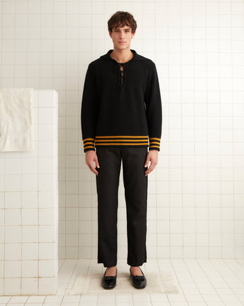 A person stands in a tiled room, wearing the Deck Stripe Sweater - Black, featuring yellow stripes and classic knitwear details.