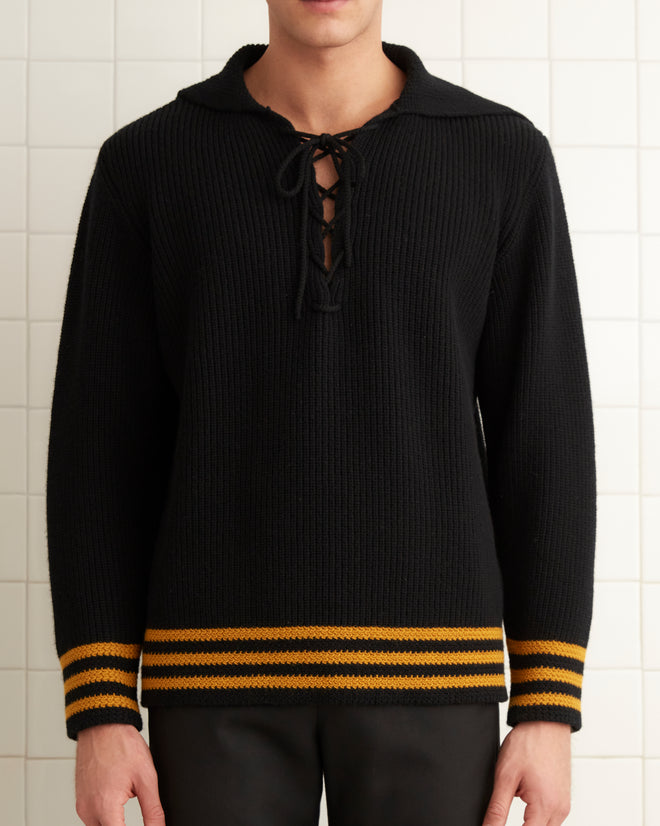 A person stands in a tiled room, wearing the Deck Stripe Sweater - Black, featuring yellow stripes and classic knitwear details.