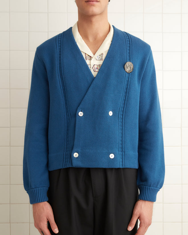 A person wearing a Double-Breasted Cardigan - Blue in a 1960s style, paired with a white shirt, black wide-leg pants, and black shoes stands in a white tiled room.