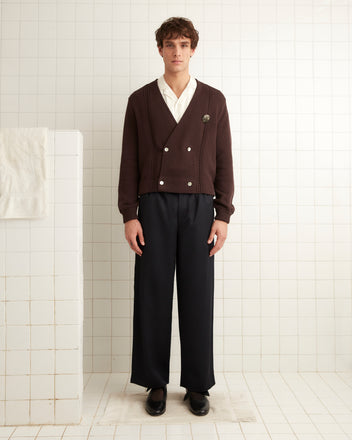 In a tiled room, an individual is dressed in a 1960s-inspired ensemble that features the Double-Breasted Cardigan in dark brown from Bode, its intricate hand-embroidered details beautifully complementing a white shirt and black wide-leg pants.