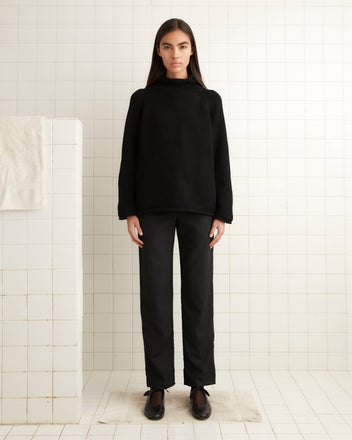 A person stands in front of a white tiled wall, wearing an oversized black Cashmere Rollneck, black pants, and black shoes. A white towel hangs on a nearby partition.