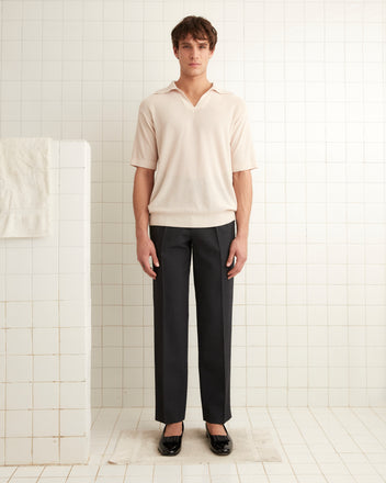 A man stands in a tiled room wearing a Boxy Polo - Ecru shirt made of light crepe yarn and dark pants. There is a wall on the left with a towel hanging on it.