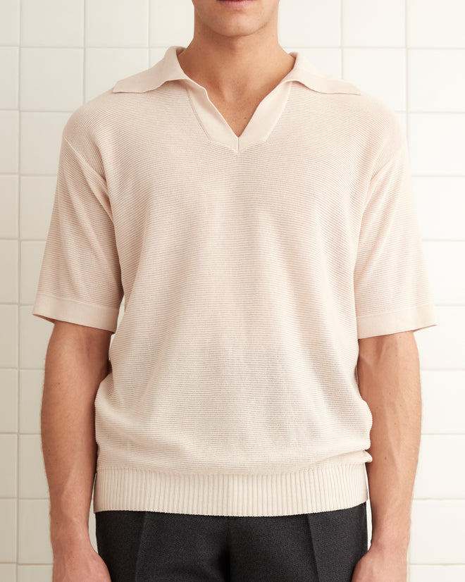 A man stands in a tiled room wearing a Boxy Polo - Ecru shirt made of light crepe yarn and dark pants. There is a wall on the left with a towel hanging on it.