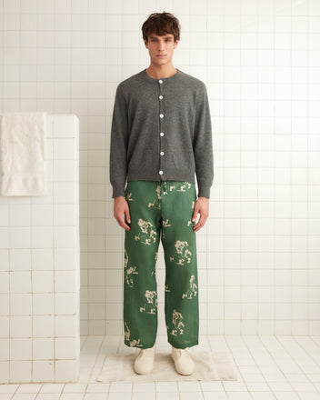 A person wearing an Orchard Cardigan in grey, featuring antique pearl buttons, along with green printed pants stands in a white tiled room.