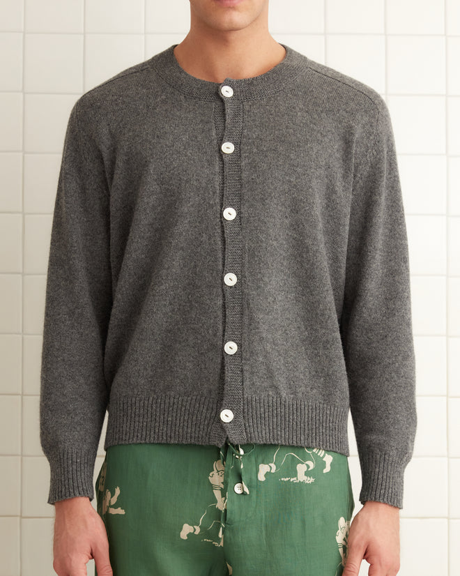 A person wearing an Orchard Cardigan in grey, featuring antique pearl buttons, along with green printed pants stands in a white tiled room.