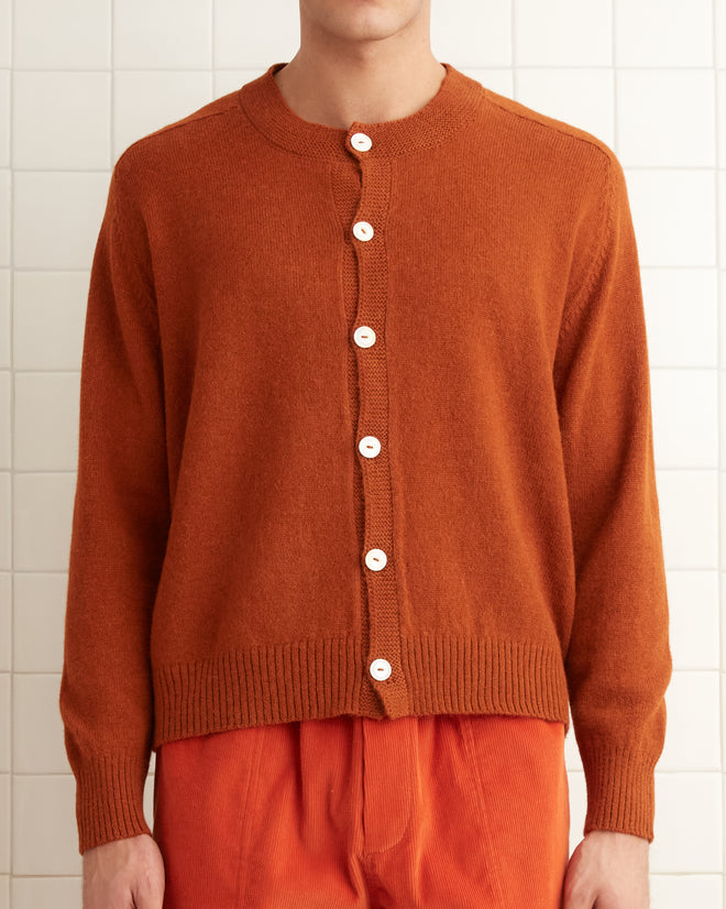 A person wearing the Orchard Cardigan in orange stands in a white-tiled room, their attire adorned with antique pearl buttons, while a towel rests neatly on the shelf.