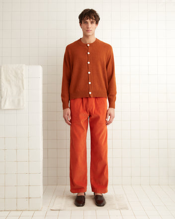 A person wearing the Orchard Cardigan in orange stands in a white-tiled room, their attire adorned with antique pearl buttons, while a towel rests neatly on the shelf.