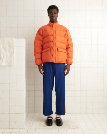 In a tiled room, a person stands wearing the Burlington Puffer Jacket in orange and blue pants, with a towel draped over a divider.