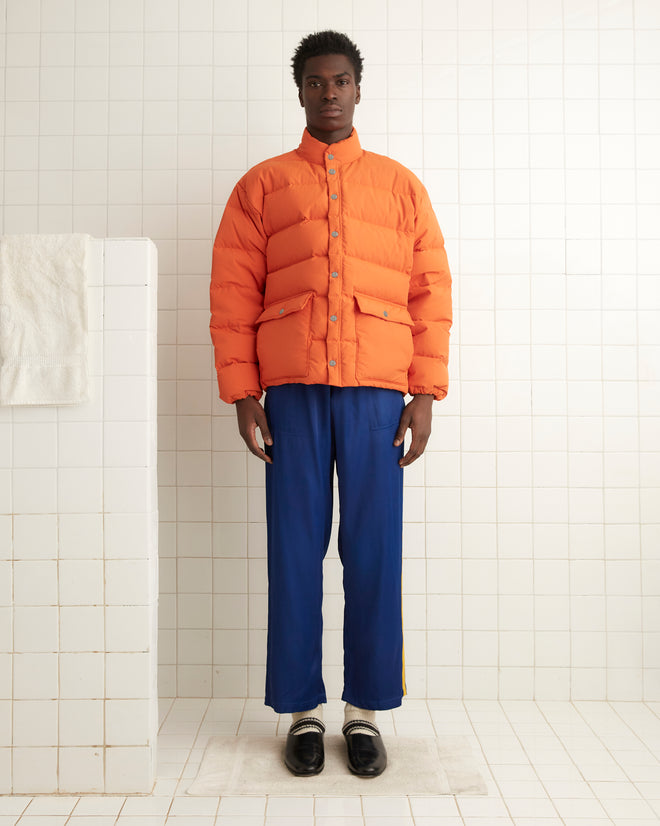 A person wearing a Burlington Puffer Jacket in orange and blue pants stands in a room with white tiles.