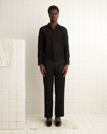 A person in a Blooming Threadwork Shirt and pants stands against a tiled wall, near a white towel crafted from 100% cotton.