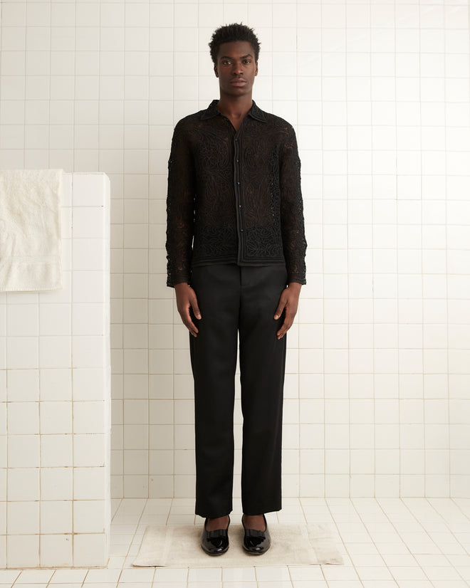 A person wearing the Blooming Threadwork Shirt and matching pants, featuring artisan handwork, stands in a tiled room with a white towel hanging on a rail nearby.