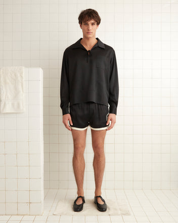 In a tiled room, an individual is wearing a black Maze Long Sleeve Shirt paired with black shorts and shoes. A folded towel is hanging on the wall to the left.