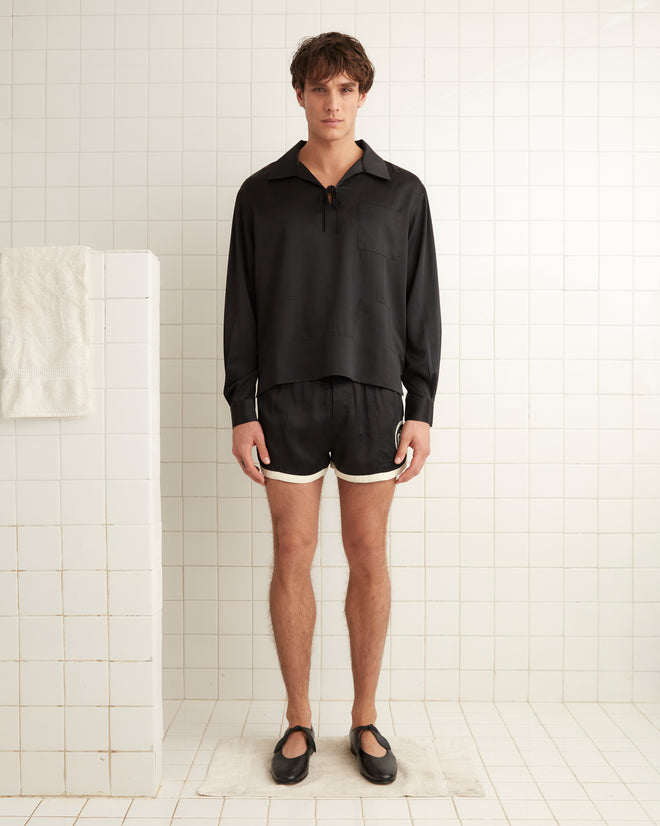 In a tiled room, an individual is wearing a black Maze Long Sleeve Shirt paired with black shorts and shoes. A folded towel is hanging on the wall to the left.