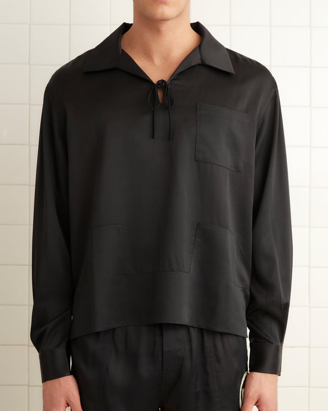 In a tiled room, an individual is wearing a black Maze Long Sleeve Shirt paired with black shorts and shoes. A folded towel is hanging on the wall to the left.