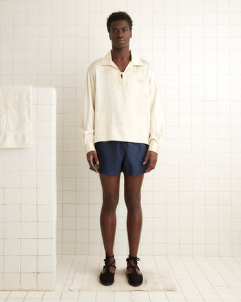 A person wearing a 1930s-inspired Maze Long Sleeve Shirt in cream and navy shorts stands in a tiled room, with a towel hanging on the wall.