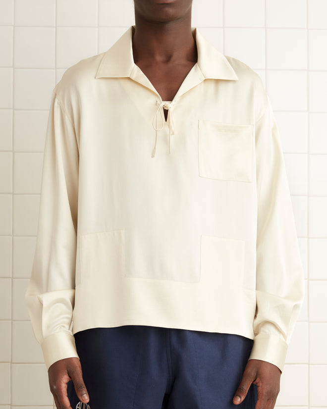 A person wearing a 1930s-inspired Maze Long Sleeve Shirt in cream and navy shorts stands in a tiled room, with a towel hanging on the wall.